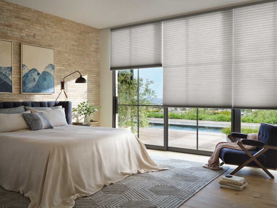 Alustra® Duette® Honeycomb Shades in a bedroom. Floor to ceiling black trim windows with a bed on the left wall with a textured wall and a modern picture over the bed. The bed has a cream draping bedspread and a pattered rug underneath. The rug is a neutral color.