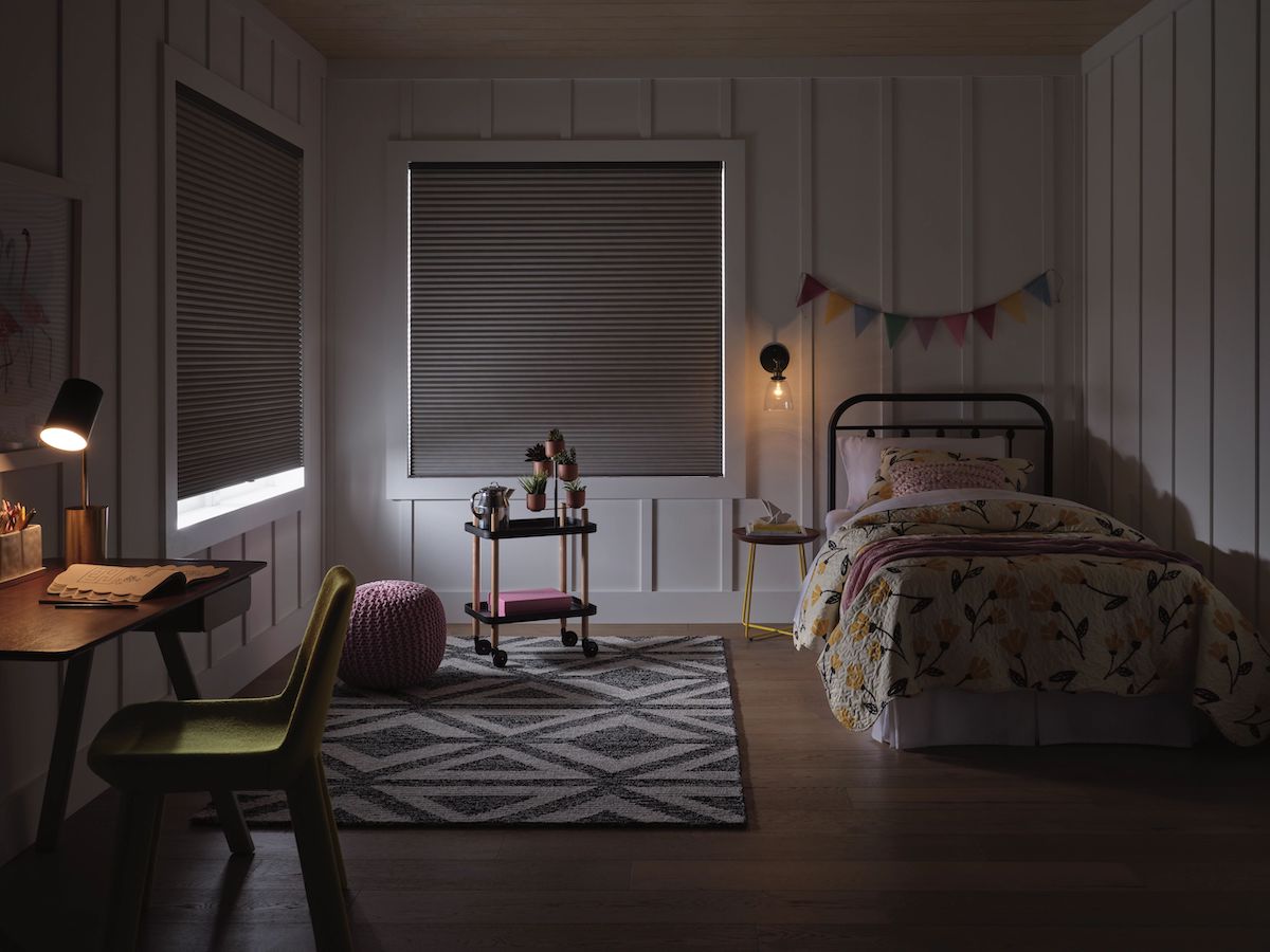 Applause® Honeycomb Shades in a dark child's bedroom. There is a twin bed against the wall on the right with two windows on the left and back wall with a peek of sunlight coming through.