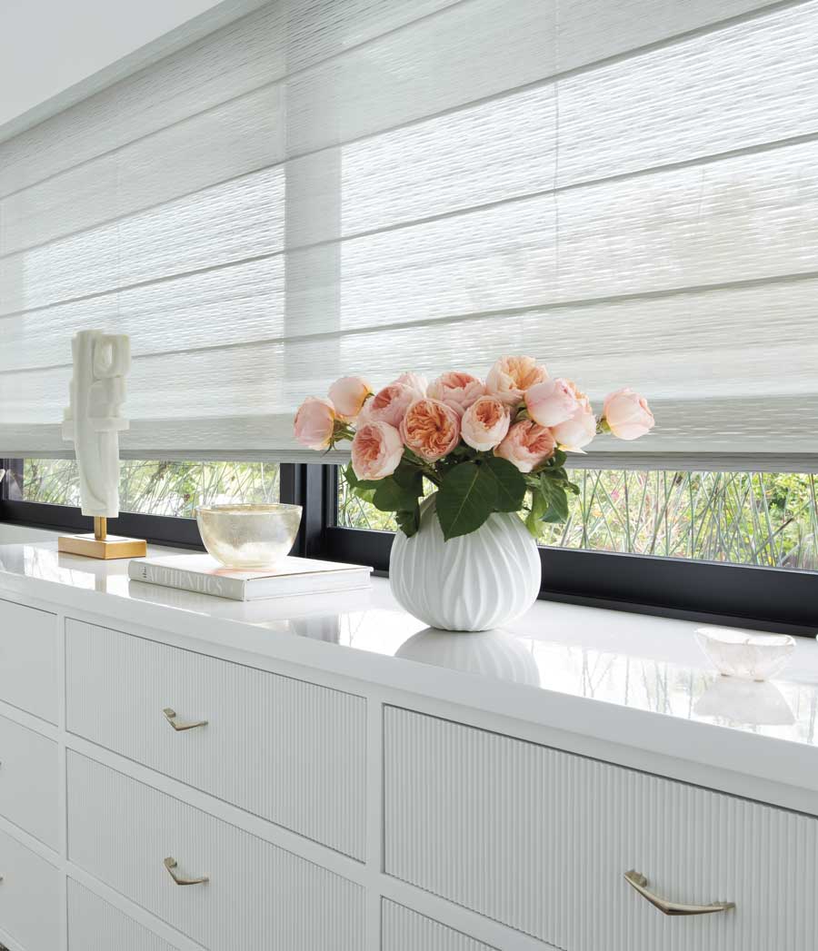 A white vase of peach colored roses sit on a white dresser with white Alustra® Woven Textures® shades