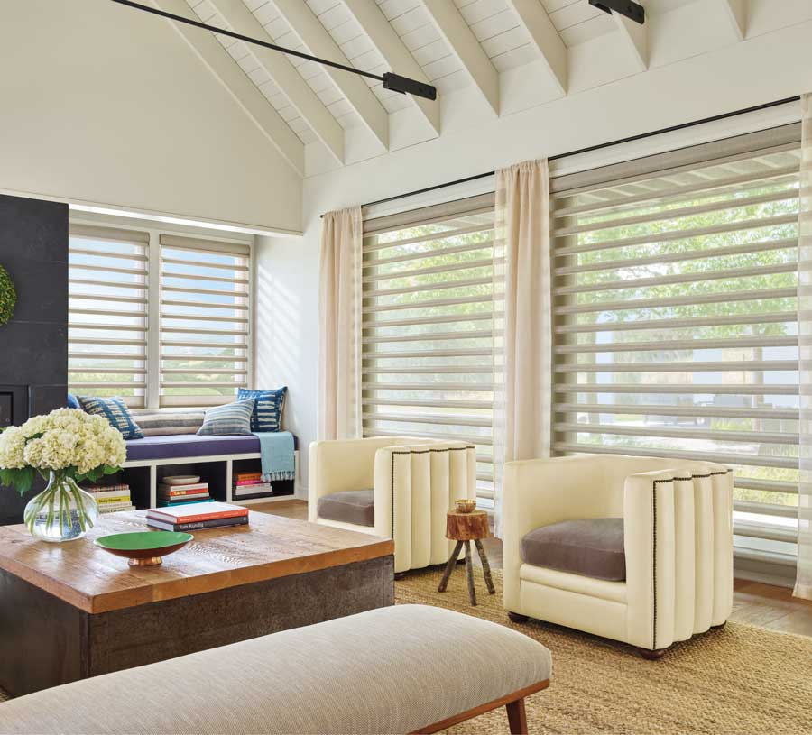 A modern living room with vaulted white ceilings and floor to ceiling windows that have drapes on either side that match the windows and pirouette shades covering the glass. The living room has two chairs, a bench and a couch all surrounding a central coffee table