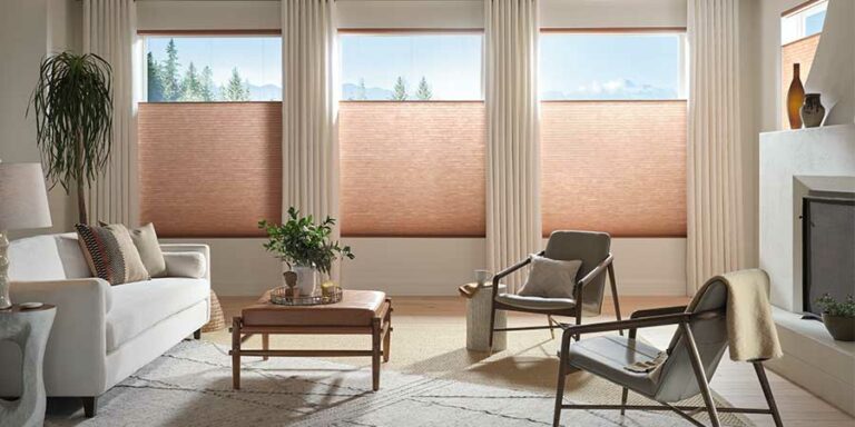 Applause® Honeycomb Shades in a big living room with exposed wooden beams, a white couch, two modern chairs and square coffee table in the middle. It is all neutral colors except the peach colored cellular shades.